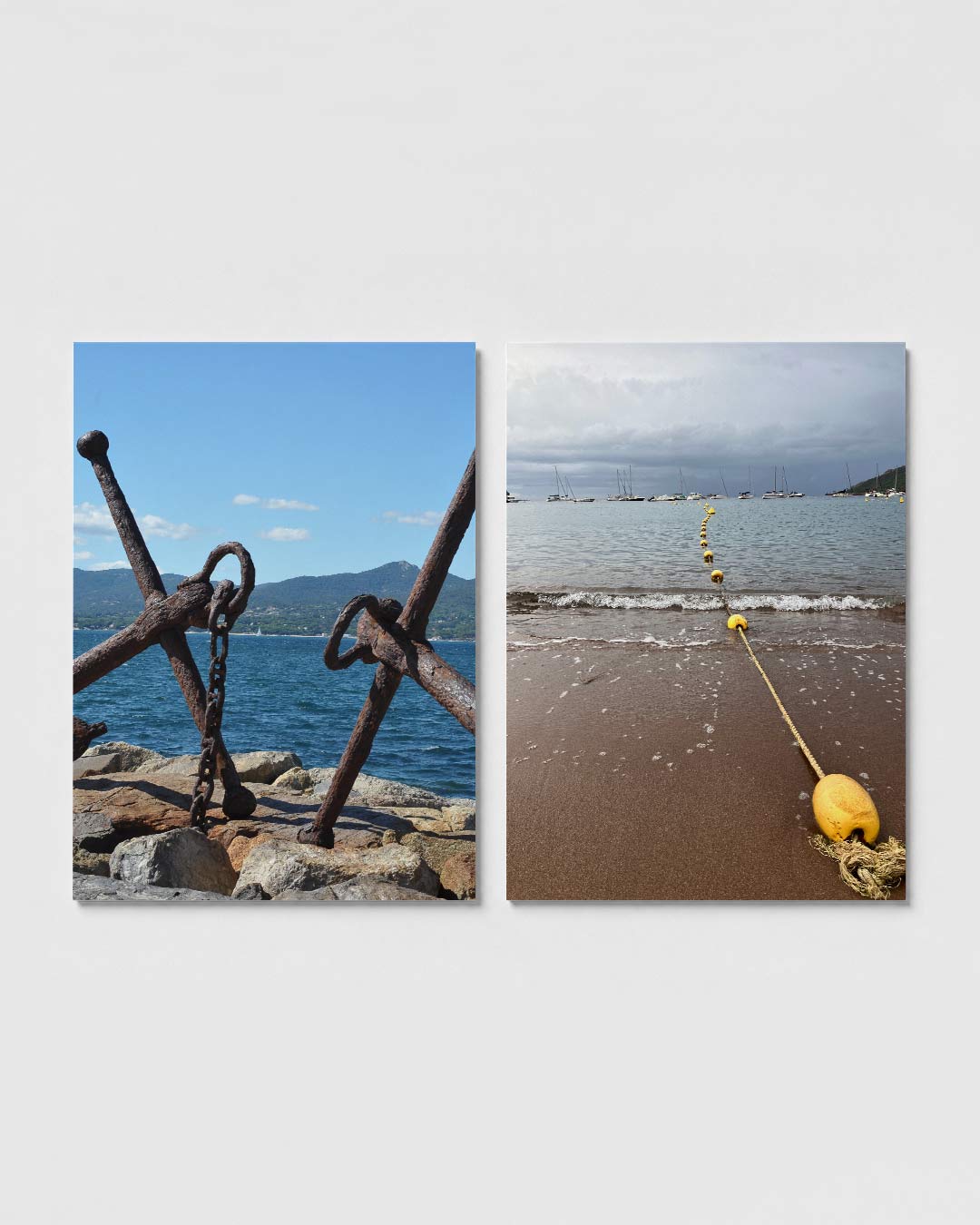 Anker en zee posterset – sfeerbeeld – twee verweerde ankers rusten op rotsen aan de kust, met een blauwe zee en heuvels op de